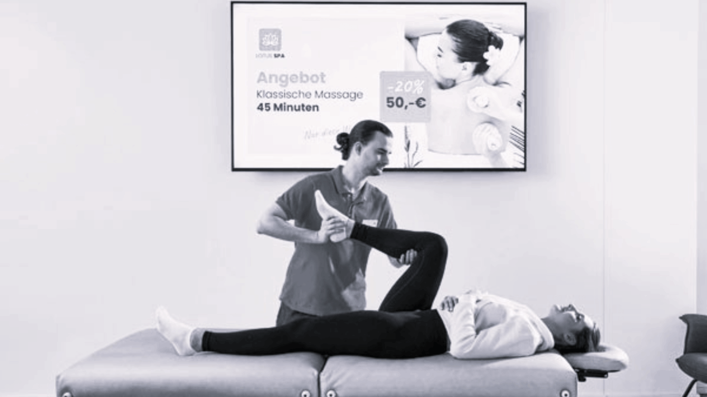 Physiotherapeut bei der Behandlung, im Hintergrund Werbebildschirm